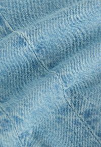 Ljusblå denim tyg med en texturerad yta, som visar ett subtilt vävningmönster och synliga sömmar. Mjuk och något urblekt framtoning.
