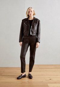 Zwarte leren cropped jas met zilveren ritsen, gecombineerd met strakke zwarte leren broeken en loafers. Houten vloer en neutrale achtergrond.