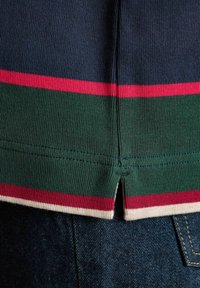 Nahaufnahme eines gestrickten Kleidungsstücks mit navy, grün, rot und weißen Streifen am Saum, mit einem Schlitz in der Mitte des Rückens über einer dunkelblauen Jeans.