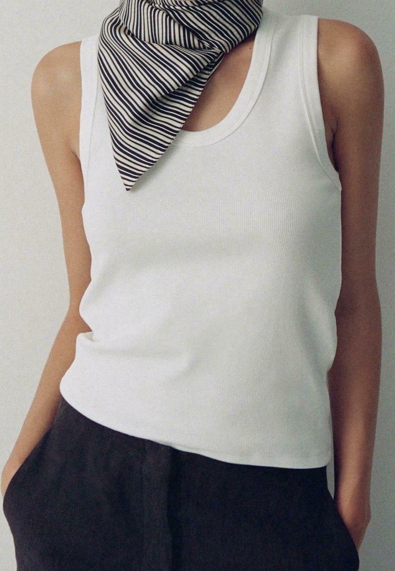 Personne portant un haut blanc sans manches, un pantalon noir et une écharpe rayée noir et blanc nouée autour du cou.