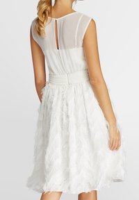Robe blanche avec un corsage ajusté et une jupe évasée à texture semblable à des plumes. Panneau arrière transparent avec un petit détail en forme de trou de serrure.