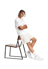 T-shirt bianco in cotone con maniche corte, pantaloni corti beige, scarpe da ginnastica bianche. Modello seduto su una sedia di metallo nero con sedile in legno.