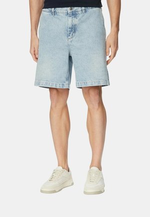 Uomo che indossa pantaloncini di jeans azzurri chiari e sneakers bianche, in piedi contro uno sfondo semplice, visibile dalla vita ai piedi.