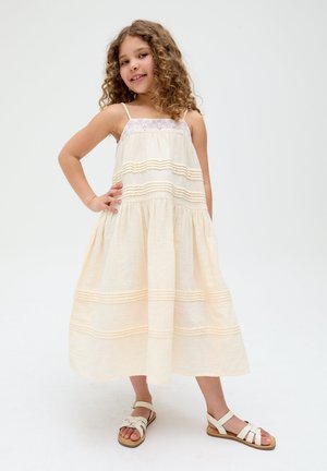 Girl with curly hair wearing a cream sleeveless dress and white sandals, standing with one hand on hip against a plain background.