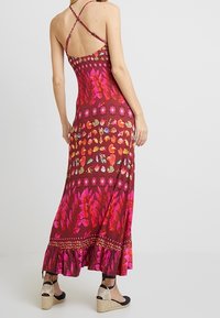 Maxi dress in burgundy, featuring multicolored floral patterns, spaghetti straps, and a ruffled hemline. Worn with espadrille sandals.