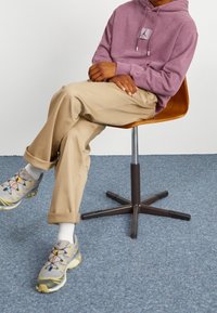 Felpa viola con logo, pantaloni beige e scarpe da ginnastica grigie con lacci colorati. Seduto su una sedia di legno contro uno sfondo chiaro.
