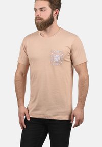 Lys beige bomull t-skjorte med korte ermer, rund hals og en mønstrete lomme med blå og oransje detaljer. Sorte jeans brukt.
