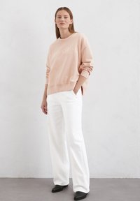 Helles rosa Sweatshirt mit besticktem "Marc O'Polo"-Logo, Rundhalsausschnitt und gerippten Bündchen, kombiniert mit weißen weiten Hosen und schwarzen Schuhen.