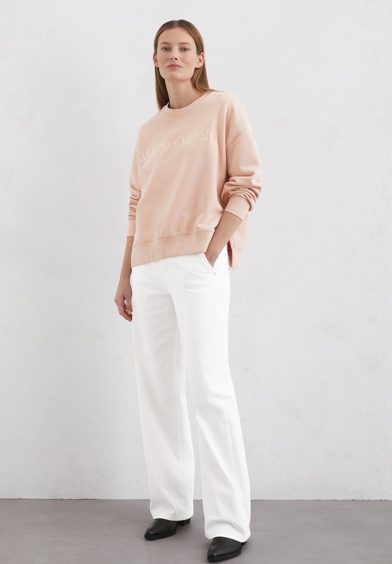 Jasnoróżowy sweter z haftowanym logo "Marc O'Polo", okrągły dekolt i ściągacze na mankietach, w połączeniu z białymi szerokimi spodniami i czarnymi butami.