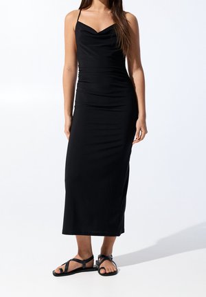 Femme portant une robe midi noire ajustée sans manches avec de fines bretelles et des sandales plates noires, debout devant un fond blanc uni.