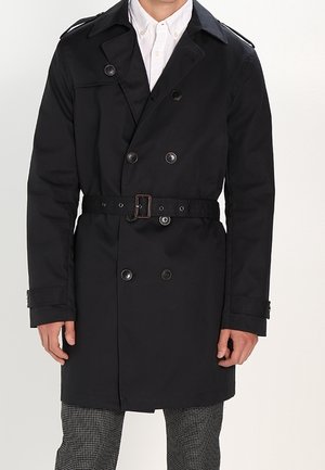 Homme portant un trench-coat noir à double boutonnage avec une ceinture sur une chemise blanche et un pantalon à motifs, debout devant un fond uni.
