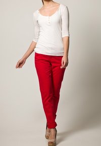 Biała koszula z długim rękawem z guzikami, zestawiona z czerwonymi dopasowanymi spodniami i beżowymi butami na wysokim obcasie, stojąca na neutralnym tle.