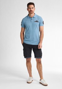 Ljusblå pikétröja med krage, korta ärmar och en patch på bröstet, tillsammans med svarta cargoshorts med uppvikta kanter och vita sneakers.