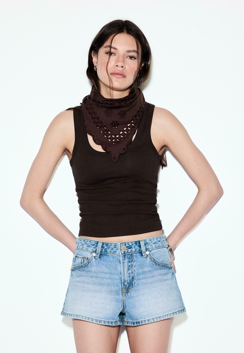 Mladá žena s tmavými vlasmi, oblečená v čiernom tielku bez rukávov, hnedom šále s vzorom a svetlomodrých džínových šortkách, stojaca s rukami za chrbtom.