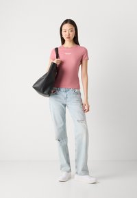 Růžové přiléhavé tričko s bílým logem, světlé distressed modré džíny a černá kabelka. Modelka v bílé obuvi stojí na neutrálním pozadí.