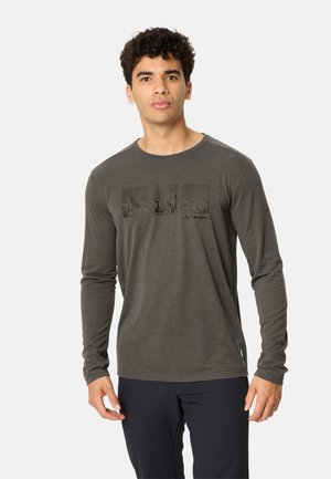 Junger Mann mit kurzen lockigen Haaren, der ein graues Langarmshirt mit dezentem Grafikmotiv und schwarze Hose trägt, steht vor einem schlichten Hintergrund.