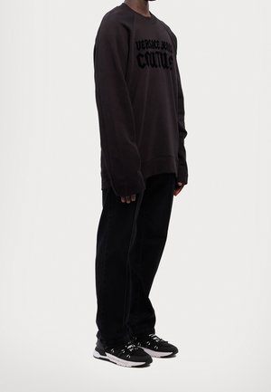 Osoba nosząca oversize czarną bluzę Versace Jeans Couture, czarne spodnie i czarno-białe trampki na jednolitym jasnym tle.