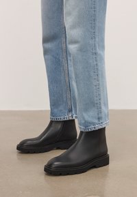 Personas, kas valkā gaiši zilos džinsus un melnas ādas potītes zābakus ar izteiktiem zoleņiem, stāv uz pulēta grīdas.