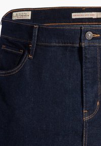Vaqueros skinny de tiro alto en mezclilla oscura con costuras naranjas, diseño de cinco bolsillos y cierre de botón metálico. Etiqueta de la marca visible en la cinturilla.