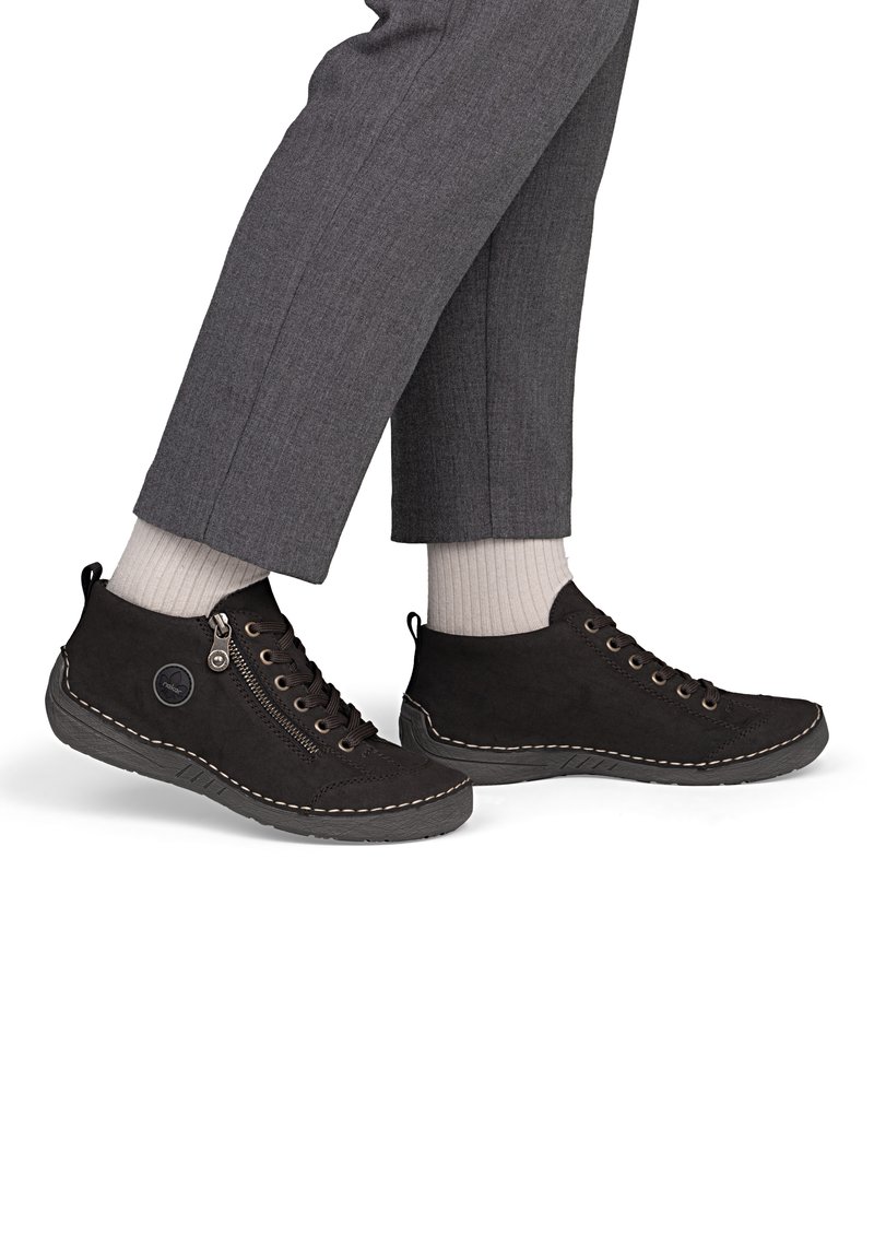 Pieds portant des chaussettes blanches et des chaussures de ville décontractées noires à lacets avec fermetures éclair latérales et semelles cousues, associées à un pantalon gris.