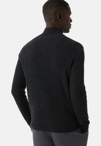 Mężczyzna w ciemnoszarym swetrze z prążkowanego dzianiny z kołnierzem, pokazany z tyłu z rękoma w kieszeniach na jednolitym tle.