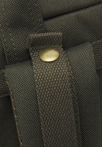 Bretelle de sac à dos en tissu texturé vert foncé avec un détail de fermeture par bouton-pression en laiton, montrant un tissage et une couture serrés.