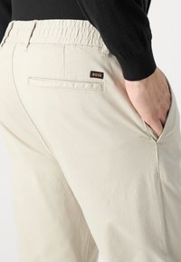 Pantalones de color beige claro con una tela texturada, cinturilla elástica, bolsillos laterales y una pequeña etiqueta negra y naranja de "BOSS" en la parte trasera.