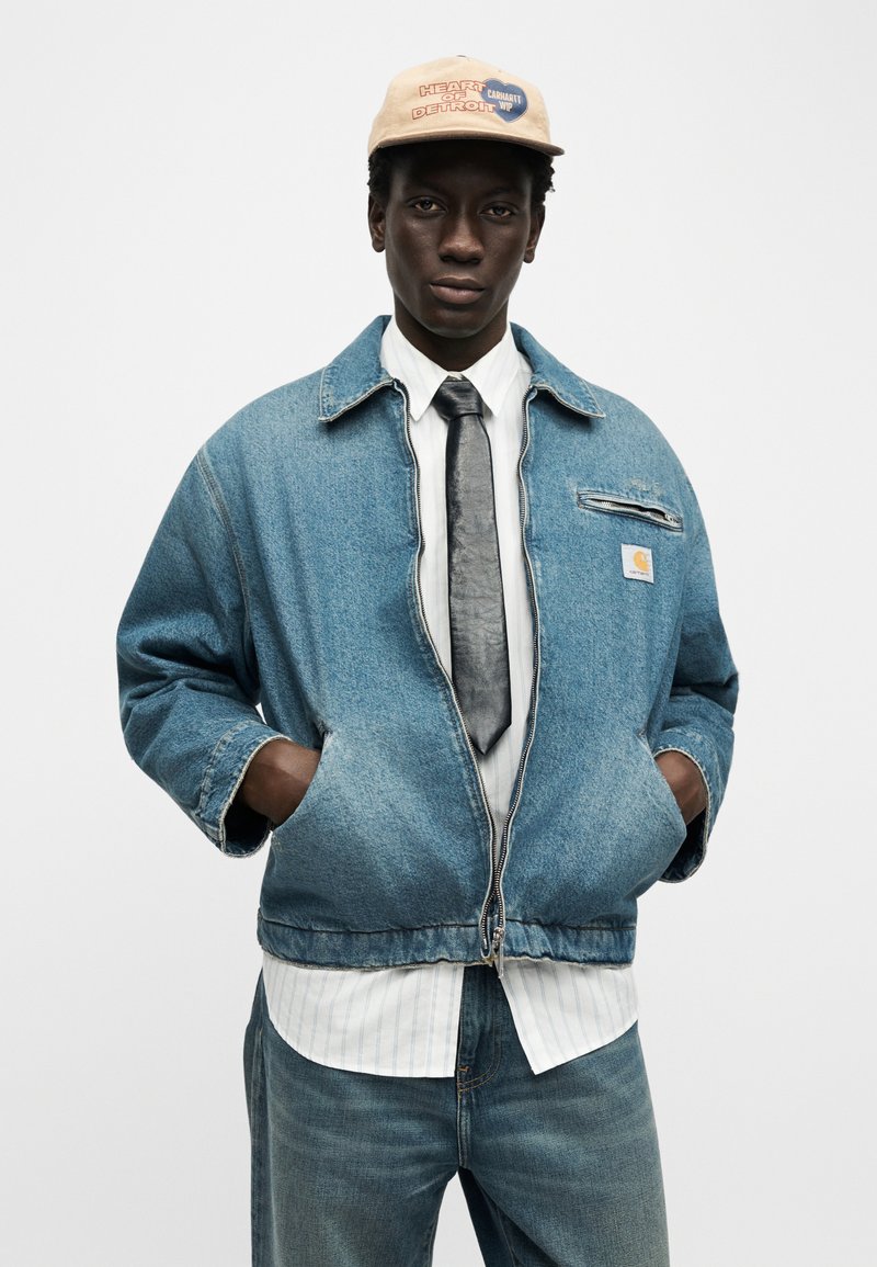 Jeune homme portant une casquette beige, une veste en jean bleue surdimensionnée, une chemise blanche à rayures verticales, une cravate noire et un jean bleu, les mains dans les poches de la veste.