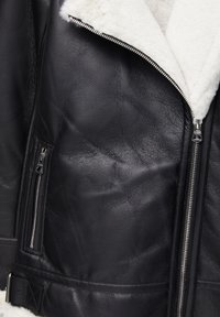Veste en cuir noir avec fermetures éclair argentées, boucle de ceinture et doublure de col en shearling blanc et doux, montrée en gros plan.
