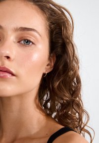 Primer plano de un rostro con cabello marrón ondulado, piel clara y maquillaje sutil. Presenta un pequeño pendiente de aro dorado y una correa negra. El fondo es claro.