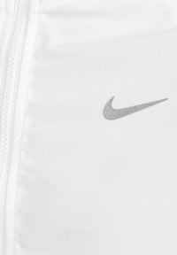 Chaqueta deportiva blanca hecha de una tela ligera con un acabado texturizado, que presenta un logotipo plateado de Nike en la parte superior derecha y un cierre de cremallera.