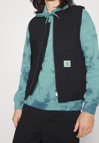 Chaleco negro sin mangas con un bolsillo frontal, usado sobre una sudadera con capucha teñida en tonos teal. Cuenta con cierre de cremallera y un pequeño parche de la marca en el pecho.