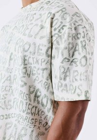 Camiseta de manga corta hecha de tela color crema que presenta un patrón de texto verde repetido. Cuello redondo con corte holgado y textura suave.
