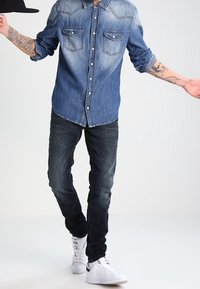 Camisa de mezclilla con tonos de azul claro y oscuro, dos bolsillos delanteros; combinada con jeans oscuros y zapatillas blancas.