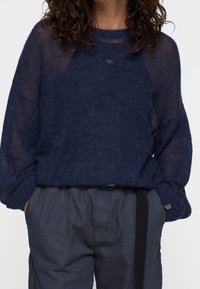 Granatowy sweter o luźnej, przejrzystej strukturze z rękawami typu raglan. W zestawie z ciemnoszarymi spodniami z kontrastującym, czarnym pasem w talii.