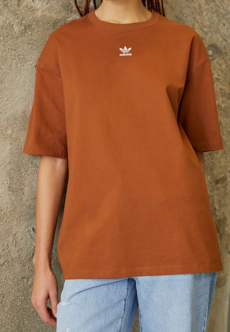 Hnědý oversized bavlněný tričko s bílým logem Adidas na hrudi. Má krátké rukávy a kulatý výstřih, kombinované se světle modrými džínami.