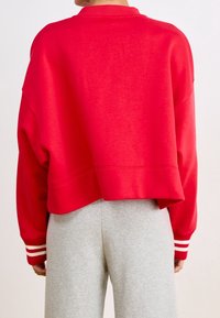 Osoba widziana z tyłu, ubrana w luźną czerwoną bluzę z białymi paskami przy mankietach oraz jasnoszare spodnie dresowe.