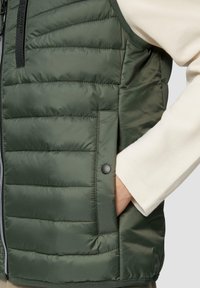 Grüne Pufferweste mit horizontaler Steppung, Reißverschluss und einer Seitentasche mit einem schwarzen Druckknopf-Detail. Weiches, glänzendes Material.