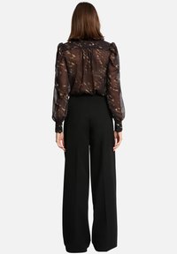 Femme vue de dos portant un chemisier noir transparent avec des accents dorés et un pantalon large taille haute noir.