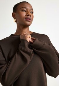 Mörkbrun överdimensionerad sweatshirt med rundad hals och breda ärmslut, med en slät yta och enkel design. Guldiga cirkulära örhängen synliga.