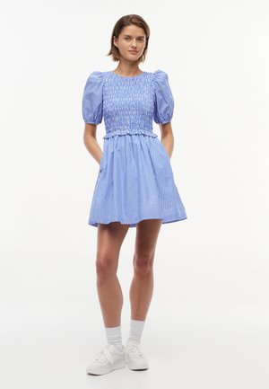 Νεαρή γυναίκα με κοντό ριγέ μπλε και άσπρο φόρεμα με φουσκωτά μανίκια, τσέπες, λευκές κάλτσες και λευκά αθλητικά παπούτσια, στέκεται με τα χέρια στις τσέπες.