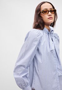 Ριγέ βαμβακερό πουκάμισο με ψηλό γιακά, λεπτομέρεια φιόγκου και φουσκωτά μανίκια. Απαλές μπλε και λευκές ρίγες, κλείσιμο μπροστά με κουμπιά, λεία υφή.