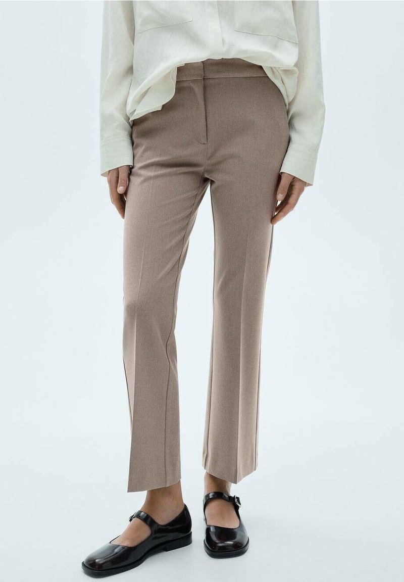 Femme portant un pantalon beige droit, une blouse blanche à manches longues et des chaussures Mary Jane noires, debout devant un fond uni.