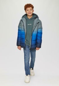 Puffer jaka ar gradienta zilu, pelēku un jūras zilu; ar kapuci un quilted tekstūru. Valkājams virs zaļas sporta krekla, kombinēts ar ziliem džinsiem un baltām kedām.