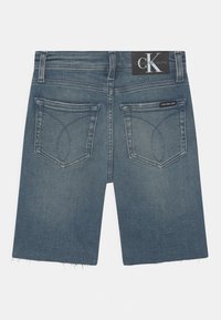 Denim shorts in medium blue with a frayed hem. Features two back pockets with curved stitching and a black logo patch at the waist.