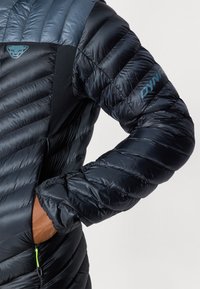Sort og blå isoleret jakke med baffle-design, sidelomme med grøn lynlås og logodetalje på skulderen. Glat, skinnende overflade.