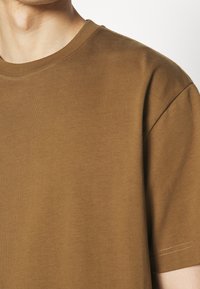 Homme portant un T-shirt simple marron à manches courtes et col rond, montré du cou au haut du bras sur un fond blanc.