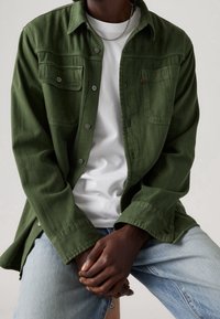 Camicia verde oliva con bottoni, due tasche sul petto, tessuto morbido, maniche lunghe e bottoni metallici, indossata sopra una t-shirt bianca e jeans in denim chiaro.