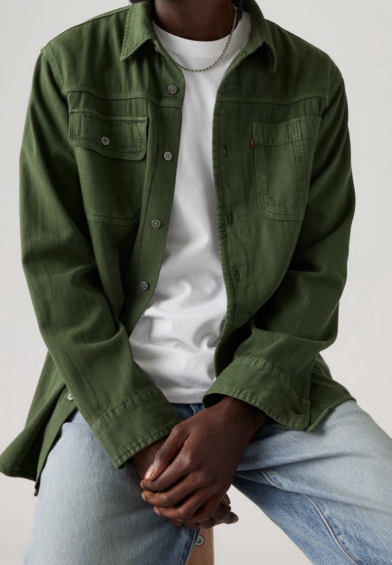 Camicia verde oliva con bottoni, due tasche sul petto, tessuto morbido, maniche lunghe e bottoni metallici, indossata sopra una t-shirt bianca e jeans in denim chiaro.