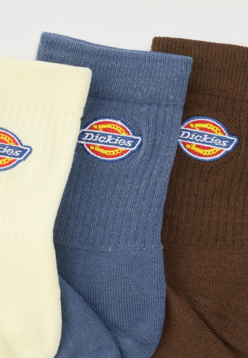 Tre calzini a coste piegati nei colori crema, blu e marrone, ciascuno con una toppa del logo Dickies multicolore vicino alla parte superiore.
